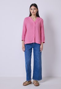 Chemisier rose à col en V, manches longues avec des détails froncés, associé à un jean bleu à jambes larges et des sandales beiges.