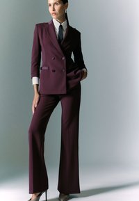 Abito su misura di un elegante bordeaux scuro con giacca doppiopetto e pantaloni a gamba larga, abbinato a una camicia bianca e una cravatta nera. Tessuto elegante e morbido.