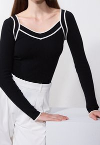 Haut noir à manches longues avec un col rond, orné de doubles rayures blanches. Porté avec un pantalon blanc taille haute, sur un fond neutre.