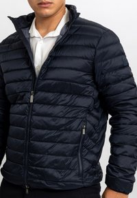 Giacca imbottita blu navy con motivo trapuntato orizzontale, collo alto, chiusura frontale con zip e tasche laterali. Tessuto liscio e lucido.