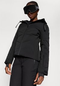 Melna pūkainā jaka ar teksturētu virsmu un rāvējslēdzēja aizdari. Iezīmējas ar kapuci, regulējamiem aproču galiem un ir saskaņota ar melniem biksēm un brillēm.