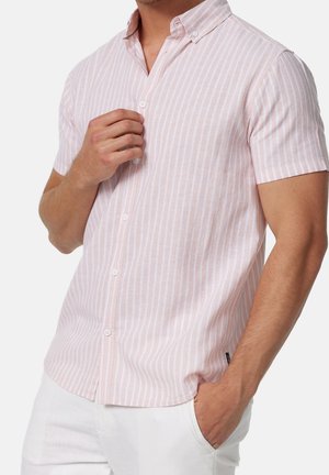 Mand iført en kortærmet, lyserød og hvid, lodret striped knap-op skjorte med den ene hånd, der justerer knapperne, og den anden i lommen.