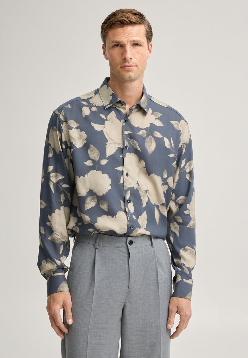 Mann steht im neutralen Licht, trägt ein blaues Hemd mit großem beigen Blumenmuster und graue plissierte Hose, blickt nach vorn.