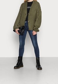 Person, die eine übergroße olivgrüne Strickjacke, ein schwarzes Graphic-T-Shirt, enge blaue Jeans und schwarze Schnürstiefel trägt, hält eine schwarze geraffte Clutch.