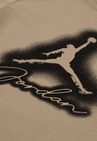 Béžový textil s texturovaným povrchem, na kterém je černá silueta skákající postavy a slovo "Jordan" v stylizovaném písmu pod ní.