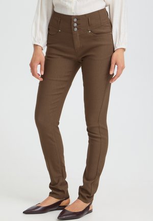 Femme portant un pantalon marron slim avec trois boutons à l'avant et des chaussures plates foncées, debout devant un fond clair uni.