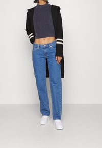 Schwarze lange Strickjacke mit weißen Streifen, graues geripptes Crop-Top, blaue gerade Jeans und weiße Sneaker vor einem einfarbigen Hintergrund.