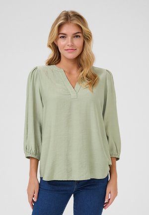 Light green blouse with a V-neck, three-quarter puff sleeves, soft textured fabric, and a relaxed fit, tucked into blue jeans.