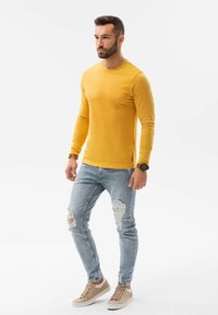 Geel langemouwen shirt, lichtblauwe distressed jeans en beige sneakers. Man staat rechtop, handen ontspannen aan de zijkanten. Eenvoudig ontwerp en pasvorm.
