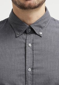 Harmaa ruudullinen paita, jossa on nappikiinnitteinen kaulus, kontrastiväriset napit ja teksturoitu kangas. Lähikuva paidan edestä.