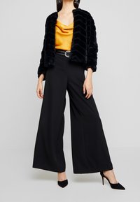 Femme portant une veste en fourrure noire, un haut en satin moutarde drapé, un pantalon noir à jambes larges, une ceinture noire cloutée et des talons pointus noirs sur fond gris.