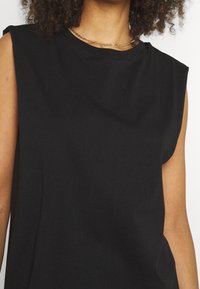 Haut noir sans manches en coton doux. Il présente un col rond et une coupe décontractée, avec une texture lisse et des épaules légèrement structurées.