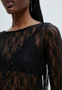 Haut en dentelle noire avec motifs floraux, détails en mesh transparent, manches longues et boutons sur le devant. Tissu doux au toucher.