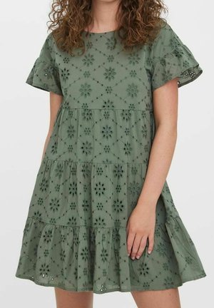 Frau trägt ein kurzärmeliges, gestuftes olivgrünes Kleid mit Lochmuster-Blumenstickerei und steht vor weißem Hintergrund.