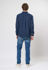 Tamsiai mėlyna marškiniai, ilgomis rankovėmis, su mygtukų rankogaliais, nugaros šoną detalė, dėvimi su šviesiai mėlynomis džinsais ir pilkais batu. Lygus audinys, pritaikyta forma.