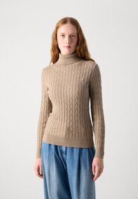 Pull en gros maillage beige à col roulé avec un ourlet et des poignets côtelés. Présente un petit logo sur la poitrine. Texture de tissu douce.