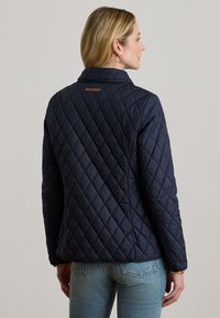 Μπλε σκούρο μπουφάν με βαμβακερή επένδυση, γιακά που στέκεται και δερμάτινη λεπτομέρεια στο πίσω μέρος. Διαθέτει διαγώνιο ράψιμο και ελαφρώς εφαρμοστή σχεδίαση.