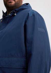 Marineblauer Windbreaker mit Kapuze, aus strukturierter Stoff, mit einer Fronttasche und einem kleinen Logo-Patch am linken Ärmel.
