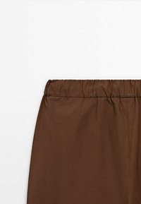 Tissu marron à ceinture élastique d’un vêtement, montrant des fronces et une texture lisse sur un fond blanc uni.