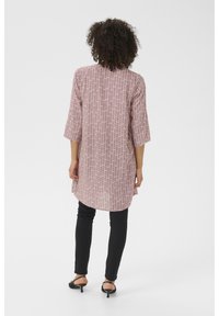 Tunique rose longue à motifs avec des manches trois-quarts, ourlet courbé et fente dans le dos. Associée à un pantalon noir ajusté et des talons ouverts.