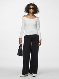 Sieviete ar cirtainiem matiem, valkājot baltu off-plecu topu, platām melnām džinsa biksēm, melnas saulesbrilles, tur mazu melnu somiņu, stāv uz balta fona.