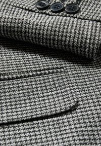 Tejido texturizado gris y negro de un abrigo con un patrón de pata de gallo, que presenta botones negros y un detalle de bolsillo.