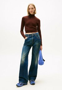 Maglia aderente a maniche lunghe marrone, jeans wide-leg blu con dettagli sbiaditi, borsa trapuntata blu e scarpe sportive blu. Sfondo bianco semplice.