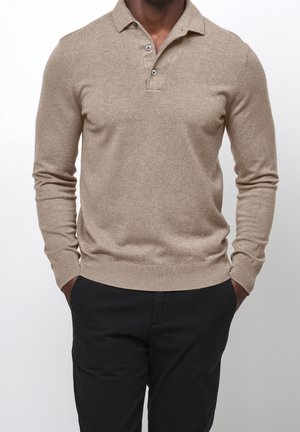 Homme portant un polo beige à manches longues avec deux boutons et un pantalon noir, les mains dans les poches, debout devant un fond clair uni.