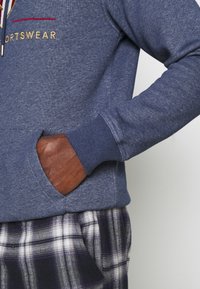 Sudadera azul de tela suave con un bolsillo delantero, puños acanalados y bordado en dorado de "SPORTSWEAR". Combinada con pantalones de cuadros.