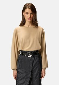 Pull beige à manches longues avec des manches raglan, associé à une ceinture noire avec une grande boucle métallique, porté sur une jupe texturée foncée.