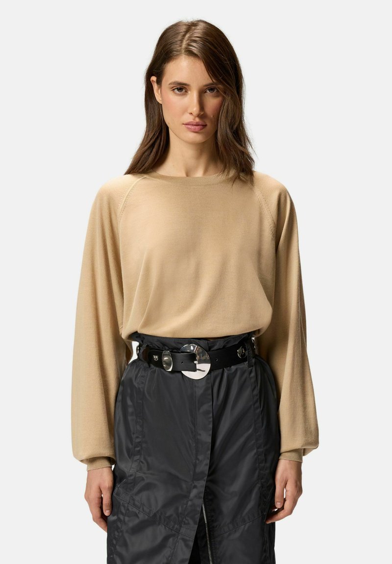 Pull beige à manches longues avec des manches raglan, associé à une ceinture noire avec une grande boucle métallique, porté sur une jupe texturée foncée.