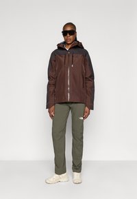 Giacca impermeabile marrone e nera con zip frontale, due tasche sul petto e cappuccio, abbinata a pantaloni verde oliva e scarpe beige.