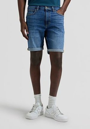 Person trägt hochgekrempelte blaue Jeansshorts, weiße Sportsocken und weiße Turnschuhe und steht vor einem schlichten Hintergrund.
