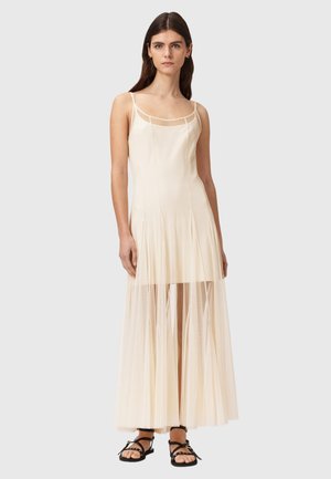 Frau trägt ein ärmelloses, cremefarbenes Kleid mit transparentem, plissiertem Überwurf und schwarzen flachen Sandalen, steht vor einem schlichten Hintergrund.