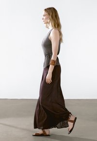Grijze mouwloze top met een wikkelontwerp, gecombineerd met een lange paarse rok en bruin platte sandalen. Kenmerkt zich door een gestructureerde stof en een brede armband.