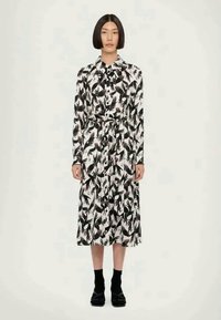 Haljina dugih rukava s crno-bijelim uzorkom lista, s pojasom u struku, midi dužine, s dugmadi sprijeda i glatkom teksturom tkanine. Nosene crne cipele.