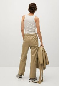 Beige neplederen broek met een elastische tailleband, gecombineerd met een witte tanktop. Een bijpassende jas hangt over één arm.