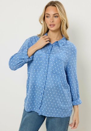 Blonde Frau trägt ein lockeres, blaues Hemd mit weißen Punkten, posiert mit einer Hand nahe am Kragen und der anderen nach unten.