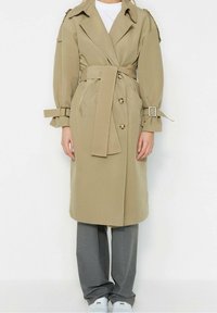 Osoba ubrana w beżowy płaszcz typu trench z paskiem i detalami guzików, szare spodnie i białe trampki, stojąca na tle jednolitej białej ściany.