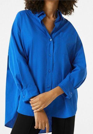 Vrouw draagt een felblauw oversized blouse met opgerolde manchetten, zwarte broek en een zilveren ketting, staand met gekruiste armen.