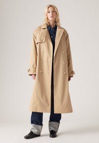 Cappotto trench beige con design doppiopetto, ampio colletto, tasche laterali e fascette regolabili sui polsini, indossato sopra pantaloni in denim scuri.