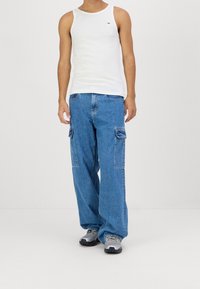 Camiseta sin mangas blanca, jeans cargo azules con bolsillos laterales, corte relajado, combinados con zapatillas grises y negras.