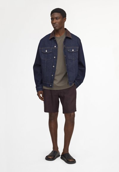 Homme portant une veste en denim foncé sur une chemise vert olive, un short noir et des sandales noires, debout devant un fond blanc uni.