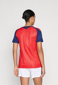 Woman wearing red and blue short-sleeve sports jersey with "España" on back collar and white athletic shorts, standing facing away.