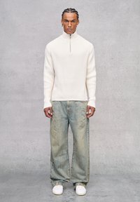 Suéter de canalé crema con media cremallera, combinado con jeans de mezclilla holgados de color azul claro y zapatillas blancas, presentando un ajuste relajado y una textura suave.