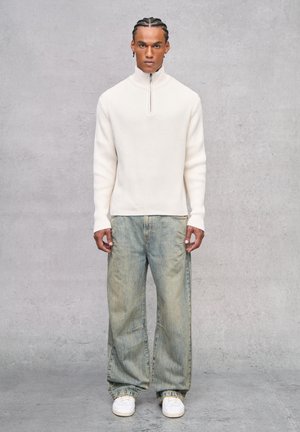Maglione a coste color crema con zip a metà, abbinato a jeans in denim baggy leggeri di colore blu e sneakers bianche, caratterizzato da una vestibilità rilassata e una texture morbida.