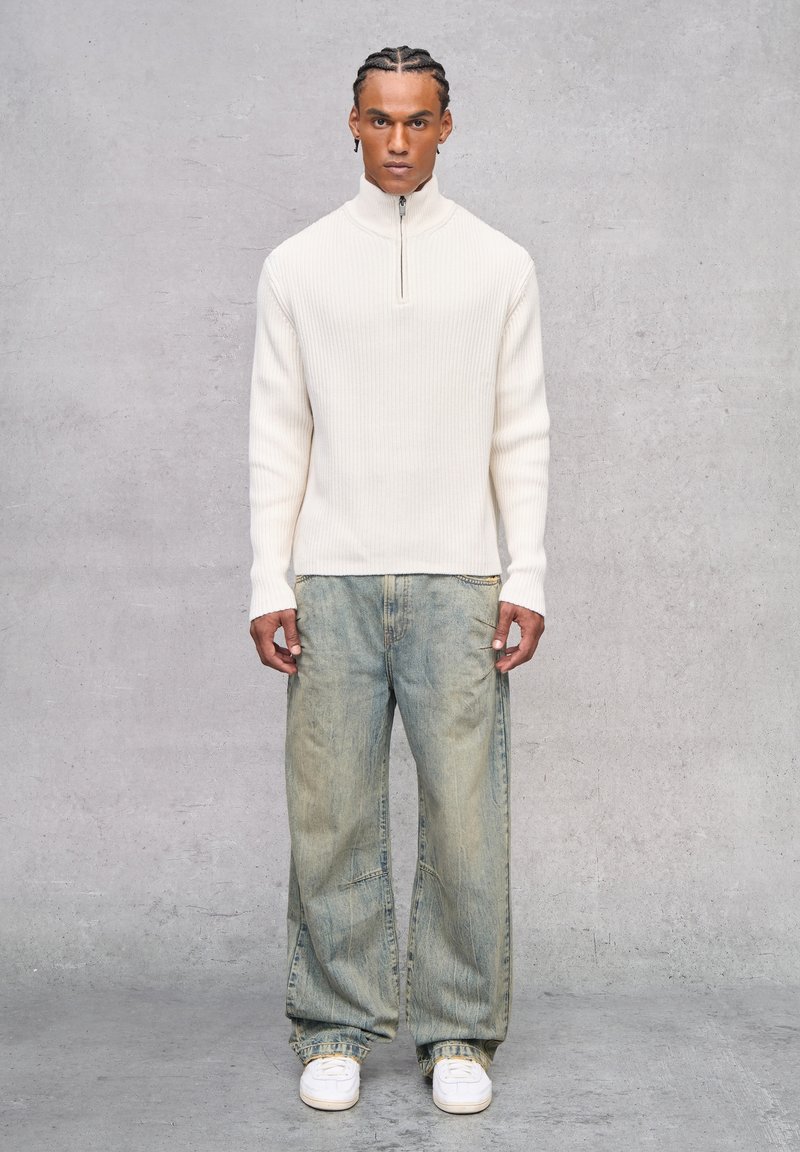 Suéter de canalé crema con media cremallera, combinado con jeans de mezclilla holgados de color azul claro y zapatillas blancas, presentando un ajuste relajado y una textura suave.
