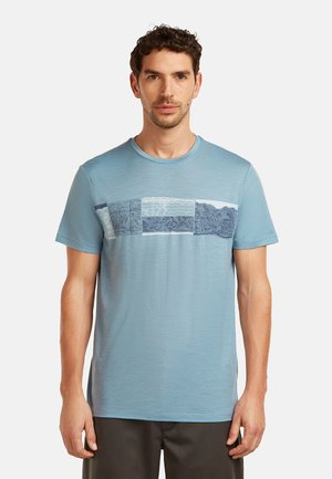 Man in een lichtblauw T-shirt met ronde hals en rechthoekige abstracte patronen op de borst, staand tegen een effen achtergrond.