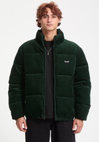 Dark green corduroy puffer jacket with a zip closure and high collar, featuring a small brand logo on the chest and a textured surface.