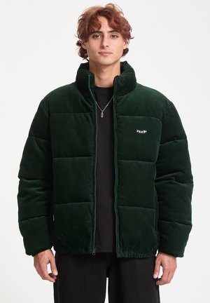 Donkergroene corduroy pufferjack met een ritssluiting en hoge kraag, voorzien van een klein merkembleem op de borst en een gestructureerd oppervlak.